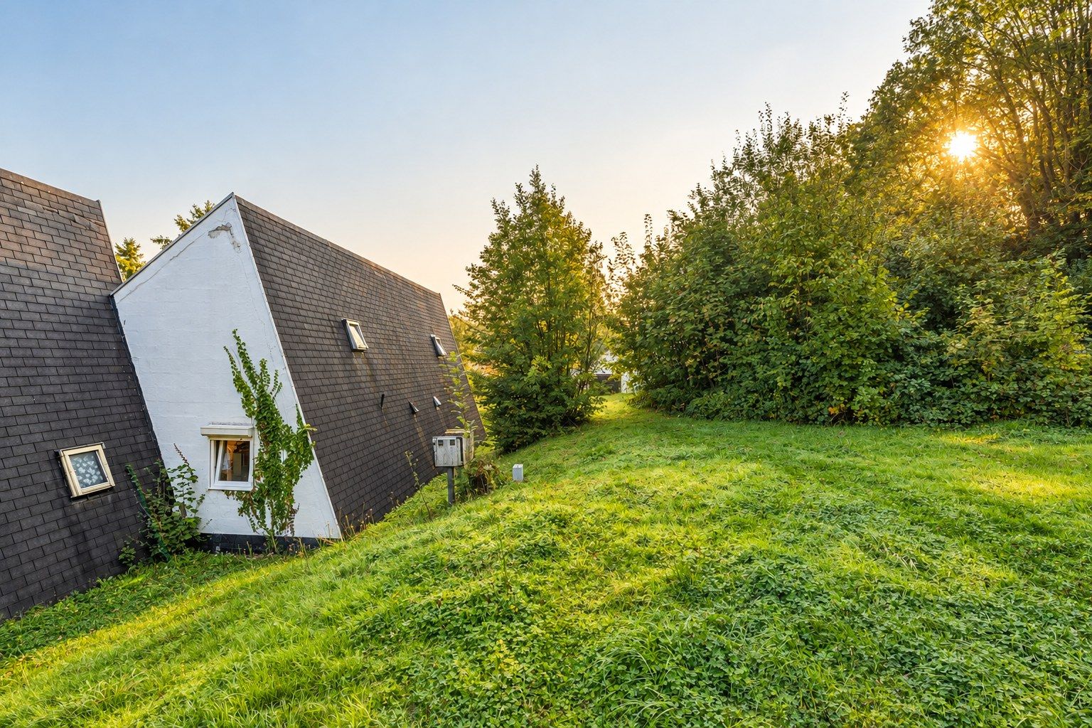 La butte herbeuse le long du pignon blanc des bungalows, bordée d’arbres, au soleil du soir