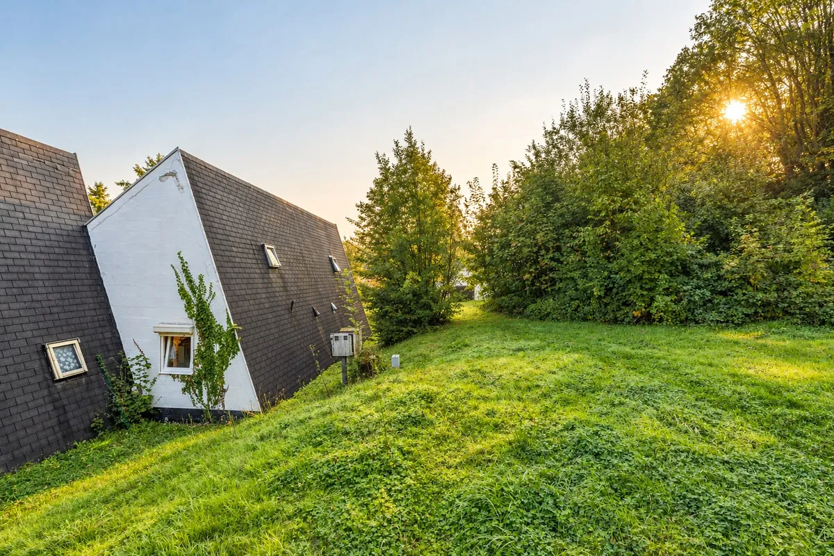 La butte herbeuse le long du pignon blanc des bungalows, bordée d’arbres, au soleil du soir
